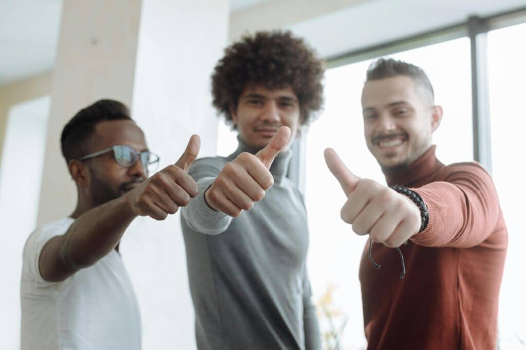 Three diverse men in a team give a thumbs up gesture indoors, symbolizing success and teamwork.